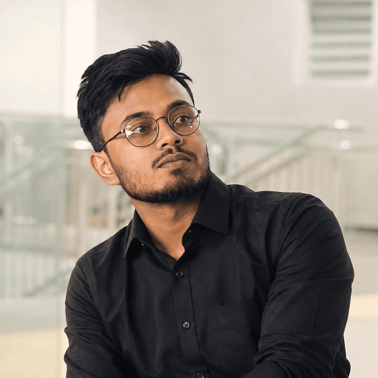 Young man with glasses and a beard wearing a black shirt, looking away thoughtfully.
